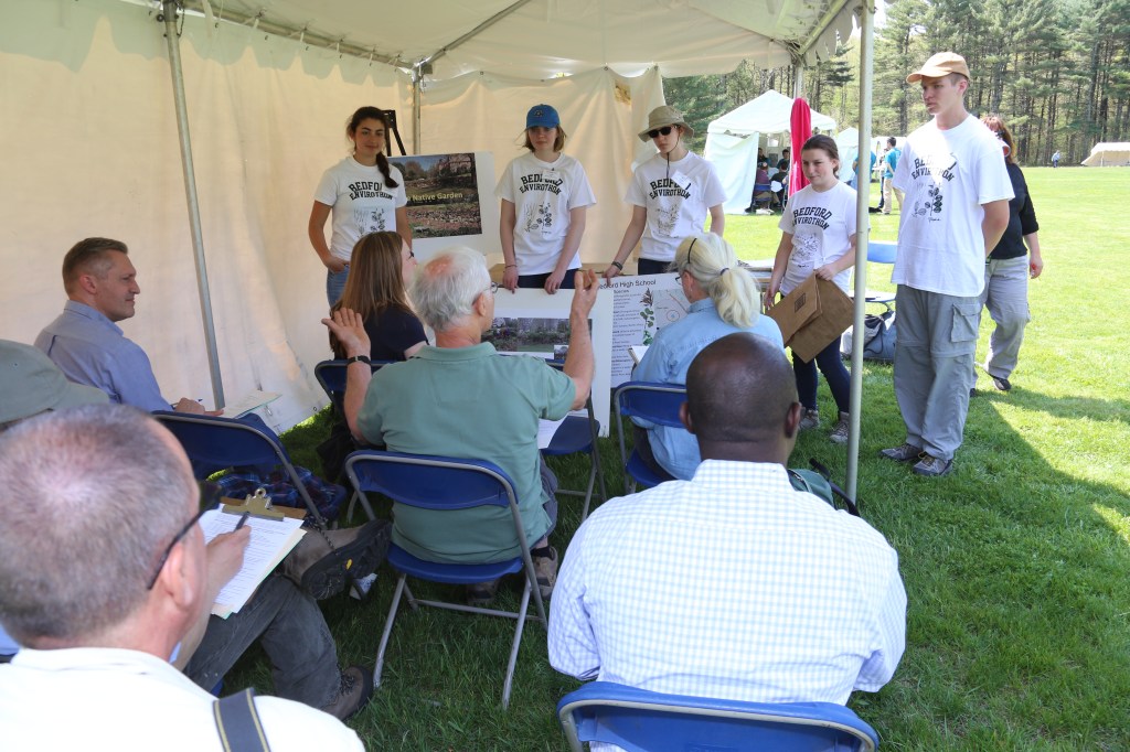 adult judges ask questions of 5 students in white t-shirts under a tent