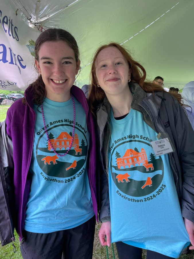 two students in light blue shirts that say oliver ames high school and show a building with wildlife, trees and a river
